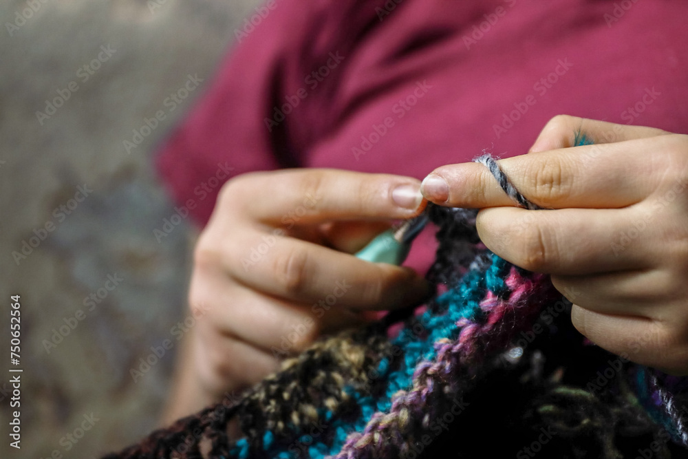 Young woman's hands crocheting colorful wool jacket with yarn sitting in the cozy arm-chair at home. Females fingers holding metal crochet hooks. Selective focus.