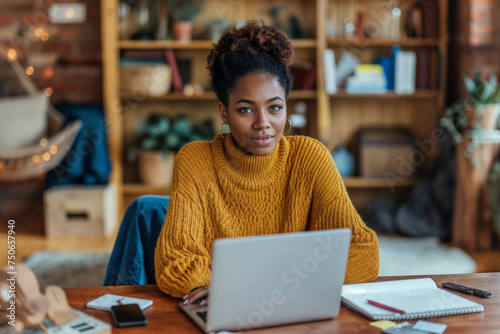 Young African American woman entrepreneur in a yellow sweater focuses on her freelance work, using a laptop in her home office