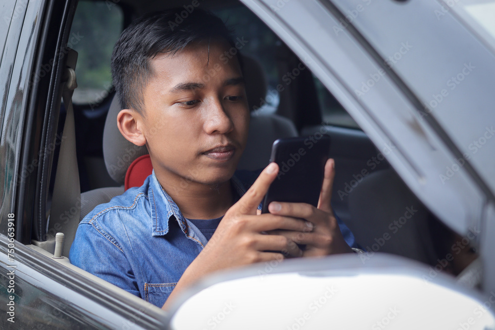 View of young Asian teenager holding and texting his phone while driving a car