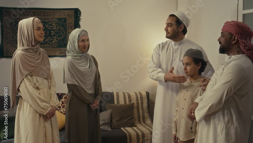 Side footage of Arab muslim man standing with son and introducing his wife and sister in hijabs to Biracial friend while gathering at home for Eid al-Fitr photo