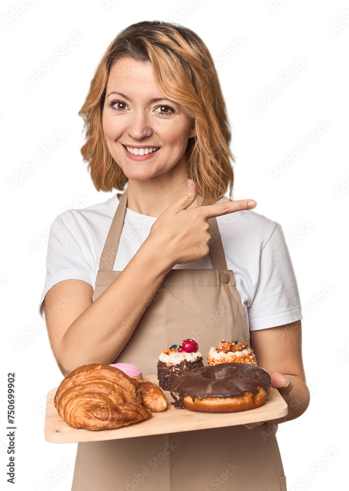 Middle-aged Caucasian woman baker with pastries smiling and pointing aside, showing something at blank space.
