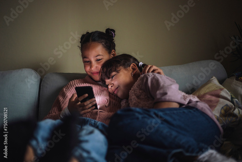Smiling female siblings sharing smart phone sitting on couch at home photo