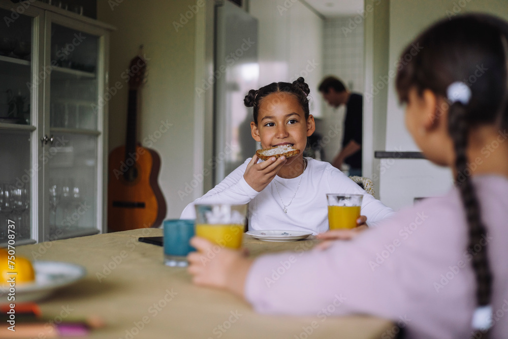 Girl eating bread slice while sitting at dining table with sister in ...