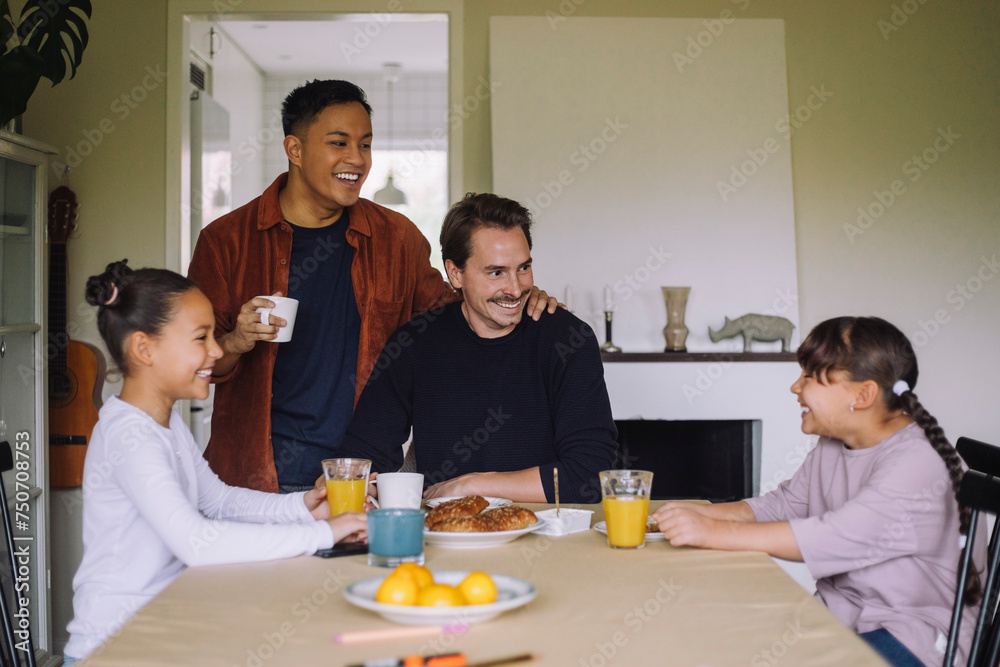Happy gay couple having fun while having breakfast with daughters at dining table in home Happy gay couple having fun while having breakfast with daughters at dining table in home