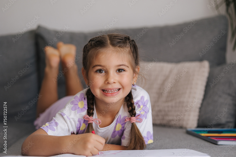 Adorable little girl with beautiful braid hairstyle lying on sofa at home and drawing picture for mom. Girl look into camera