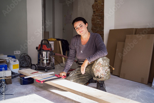 Portrait of confident female carpenter holding handsaw crouching in front of plank in apartment photo