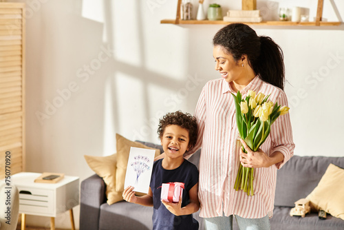 joyous african american boy looking at camera and holding postcard and present next to his mother