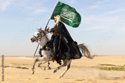 Saudi man in traditional clothing in the desert with a white horse, carrying a flag of Saudi Arabia photo