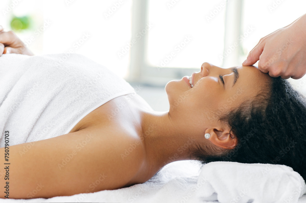 Calm relaxed brazilian or hispanic young woman lies on a massage table in a salon during a forehead massage, smiling. Female massage therapist doing general forehead massage to a pretty girl Calm relaxed brazilian or hispanic young woman lies on a massage table in a salon during a forehead massage, smiling. Female massage therapist doing general forehead massage to a pretty girl