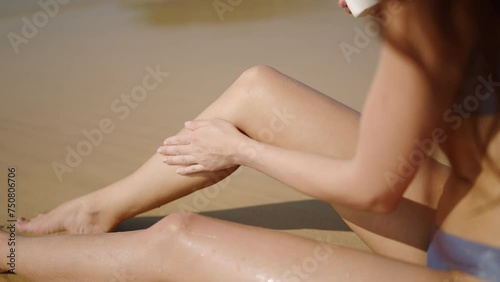 Fitted young woman applying waterproof sunscreen on legs at sandy beach. Pretty girl sits on sand with water resistant suntan lotion bottle. Tanned lady using spf. Skin protection in summer. Close-up. photo