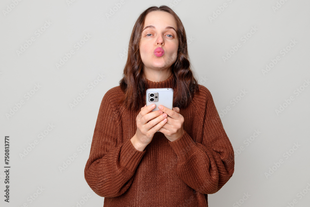 Satisfied romantic flirting Caucasian brown haired woman wearing brown jumper standing isolated over light gray background holding mobile phone sending air kissing to camera Satisfied romantic flirting Caucasian brown haired woman wearing brown jumper standing isolated over light gray background holding mobile phone sending air kissing to camera