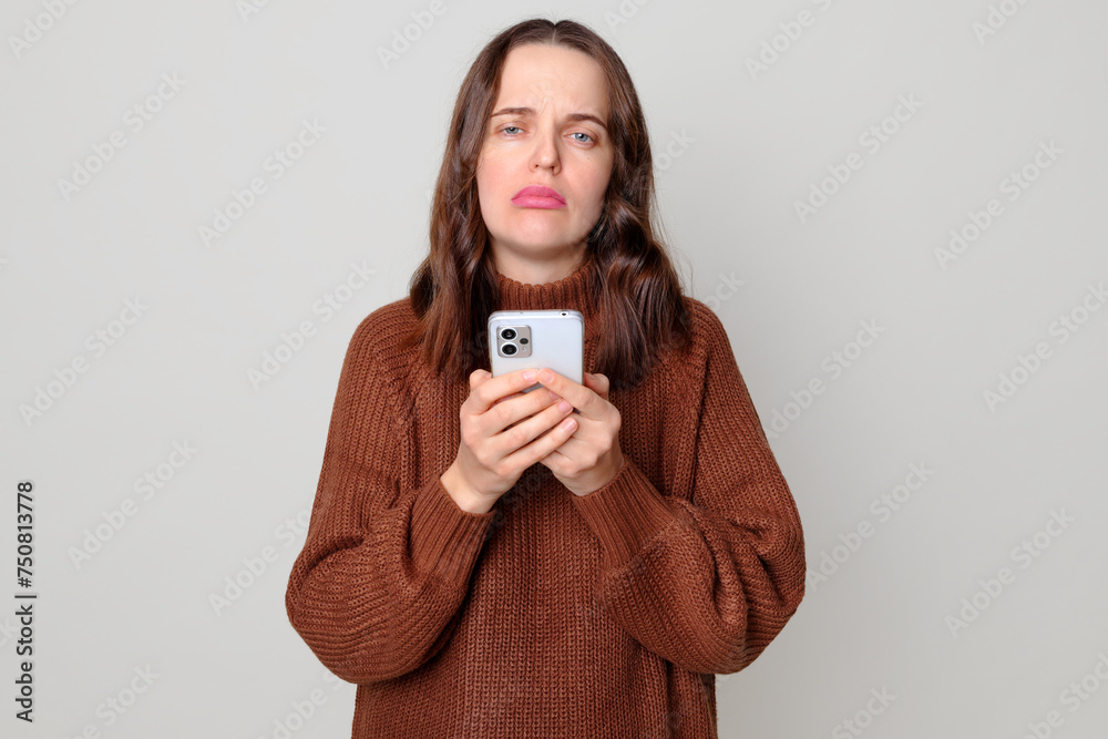 Unhappy Caucasian brown haired woman wearing brown jumper standing isolated over light gray background holding smartphone reading message with bad news Unhappy Caucasian brown haired woman wearing brown jumper standing isolated over light gray background holding smartphone reading message with bad news