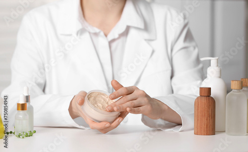 Dermatologist with jar testing cosmetic product at white table indoors, selective focus