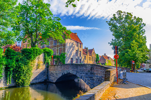 Bruges cityscape, Meebrug stone bridge across Groenerei Green water Canal with green trees and plants, embankment in Brugge old town district, medieval houses in Bruges city historic centre, Belgium