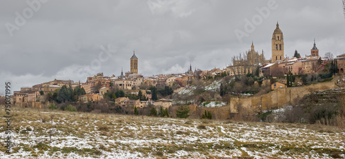 Ciudad amurallada medieval de Segovia sobre la que sobresalen la torre de la Catedral y las torres de iglesias románicas, sobre cielo gris oscuro y pradera con restos de nieve photo