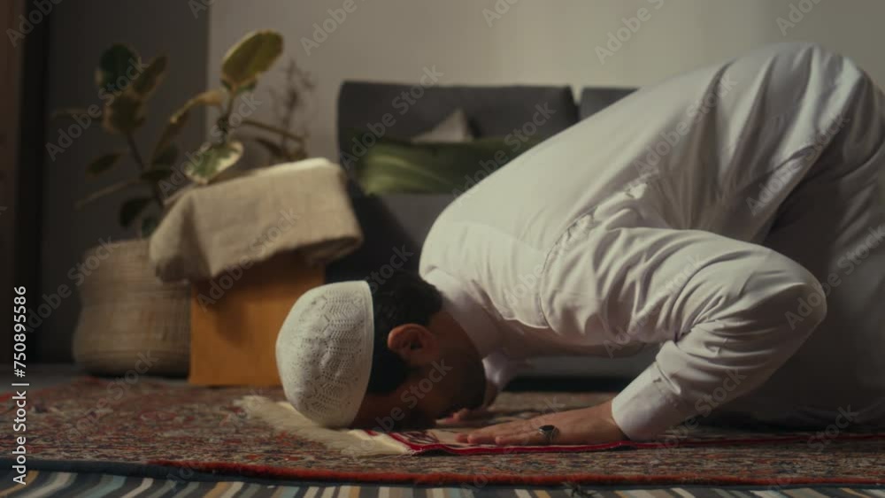 Side ground level footage of muslim man in cap and thawb prostrating while praying to Allah on prayer map at home
