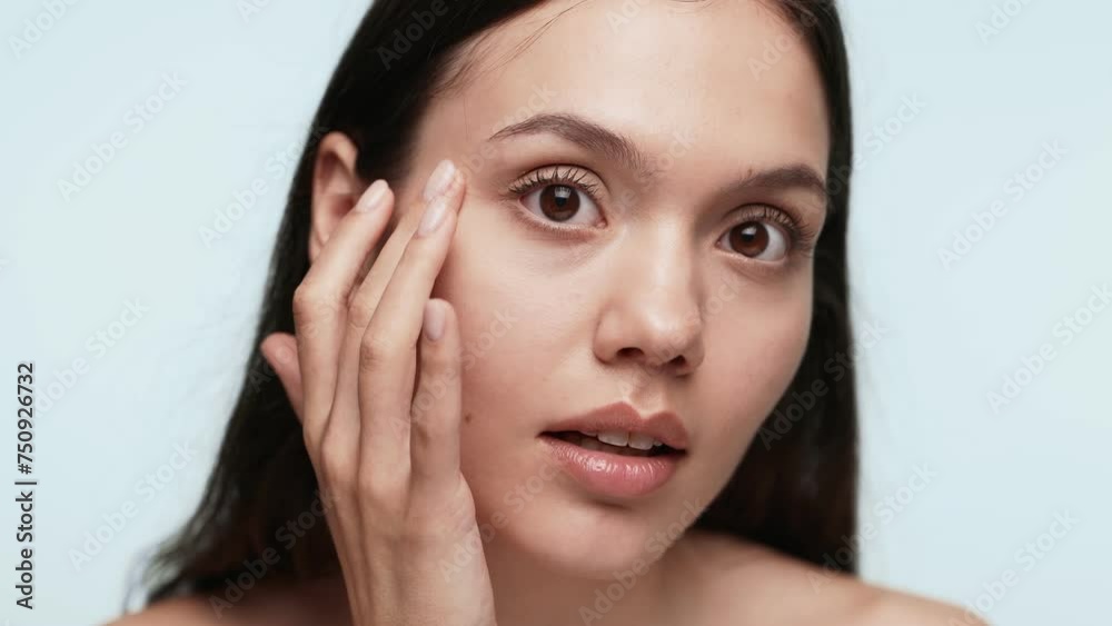 Close-up of a young woman with natural makeup touching her smooth skin on a serene blue backdrop, symbolizing skincare and beauty. Close-up of a young woman with natural makeup touching her smooth skin on a serene blue backdrop, symbolizing skincare and beauty.