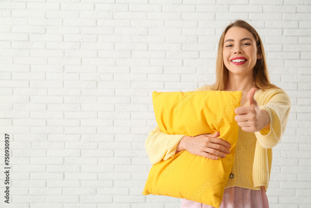 Obraz premium Beautiful young woman holding yellow soft pillow and showing thumb-up gesture on white brick background