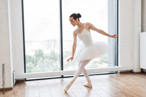 Young Caucasian ballerina in white tutu dancing on pointe in large bright modern hall in front of window. Classical ballerina. Classical ballet performed by dancer on hall of spacious studio.