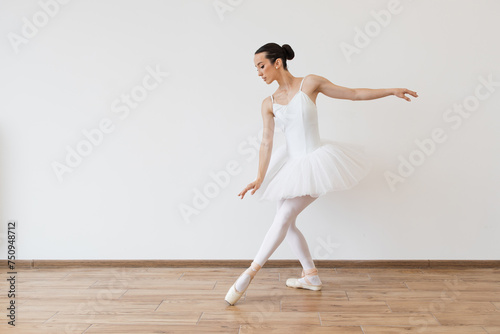 Caucasian ballerina in white bodysuit and tutu poses in motion showing ballet elements while standing on pointe shoes. Beautiful young female dancer posing on studio background.