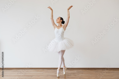 Caucasian young woman ballerina in white tutu, dancing on pointe with arms overhead, in the studio against a light bright background of modern studio hall.