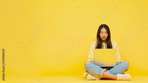 Young Asian woman working on laptop in vibrant yellow setting. Focused remote work in a casual, colorful environment.