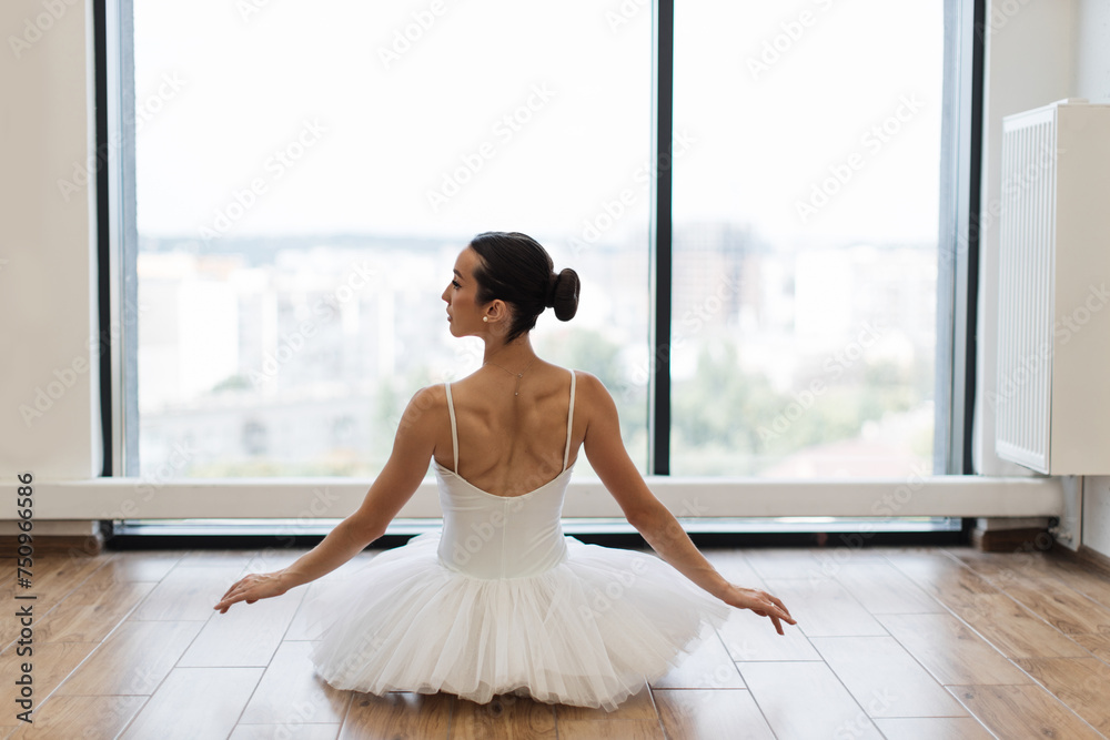 Back view of female ballet dancer sitting in front of panoramic window in natural daylight atmosphere. Focused young ballerina dressed in white tutu costume practice ballet poses at ballet studio.