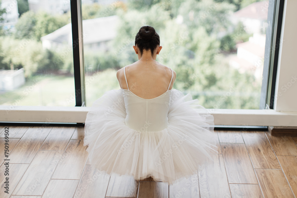 Fototapeta premium Classical Ballet dancer. Beautiful Caucasian graceful female ballerina in tutu skirt sitting at wooden floor and stretching while practice split ballet position, back view.