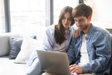 Couple in blue denim casual clothes using together a laptop sitting comfortable on the sofa at home during sunday afternoon. People working on computer at home indoor leisure online activity. Wireless