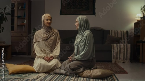 Full footage of two young muslim suisters in hijabs and abayas sitting on pillows on floor and having conversation in living room photo