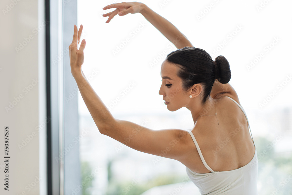 Beautiful female ballet dancer makes pirouette at ballet studio in front of big panoramic window background. Back view of focused young Caucasian ballerina dressed in white tutu costume.