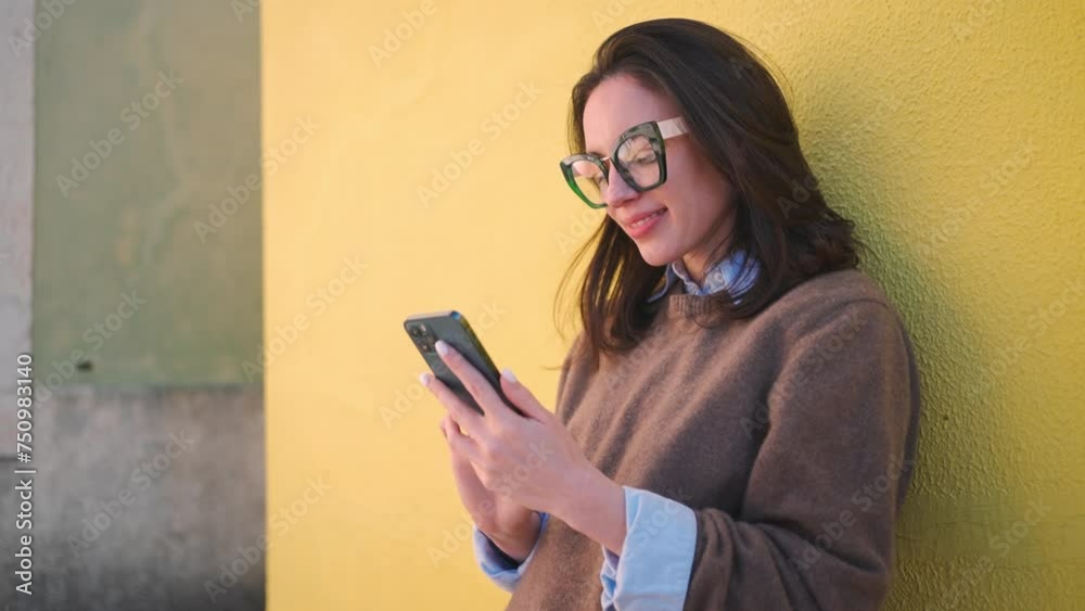Smiling woman using smartphone against yellow wall. Confident female person in smart eyeglasses and sweater text messaging via mobile phone. Lifestyle concept with cellphone. Smiling woman using smartphone against yellow wall. Confident female person in smart eyeglasses and sweater text messaging via mobile phone. Lifestyle concept with cellphone.