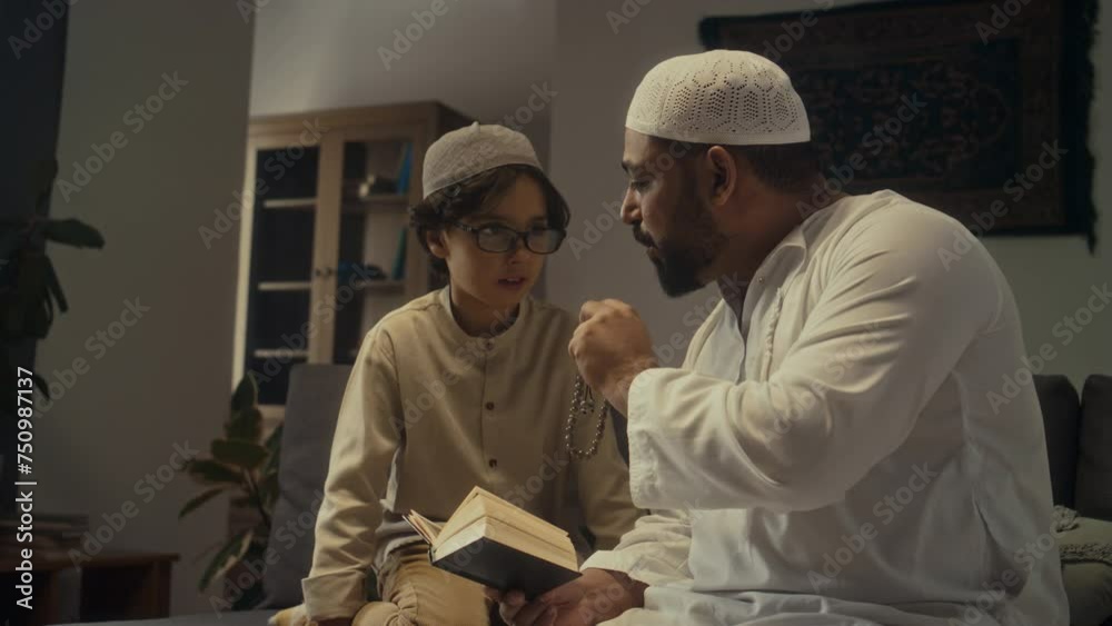 Side footage of little muslim boy in glasses attentively listening to his father in thobe and repeating arabic phrases from Quran while learning how to pray namaz using subha at home