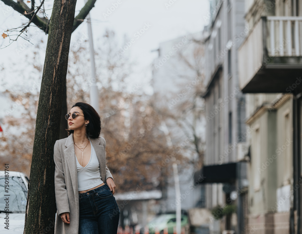 A stylish young woman in sunglasses and a chic coat stands pensively on a city sidewalk, exuding urban elegance and confidence.