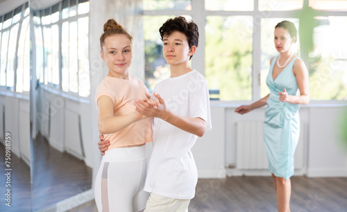 Passionate young ballroom enthusiasts, teenage girl and boy practicing graceful waltz routine in well-lit dance hall under female mentor watchful eye