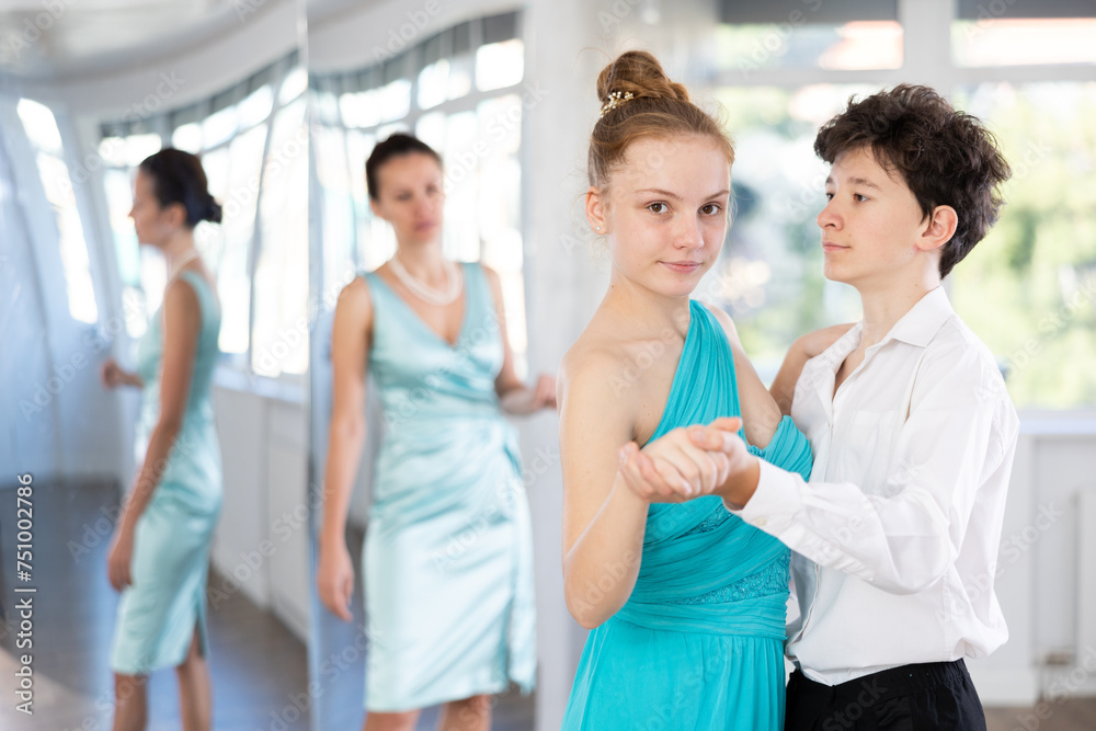 Motivated adolescent ballroom dancers, girl and boy in performance outfit practicing elegant dance moves in pair in bright studio, with female coach supervising in background