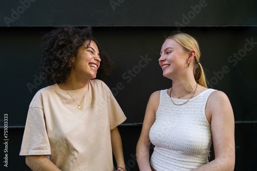 Portrait of two young diverse female friends standing against black background talking