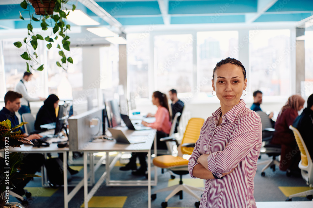 Portrait of young smiling business woman in creative open space coworking startup office. Successful businesswoman standing in office with copyspace. Coworkers working in background Portrait of young smiling business woman in creative open space coworking startup office. Successful businesswoman standing in office with copyspace. Coworkers working in background