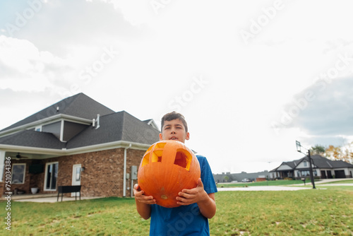 A teen boy holding a large pumpkin up with his arms.  photo