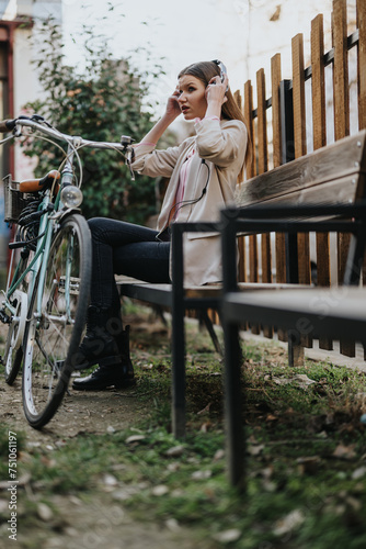 Fototapeta Naklejka Na Ścianę i Meble -  Casual young woman putting on headphones to enjoy music, sitting by a retro bicycle in an urban backyard setting.