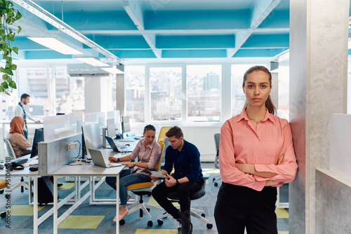 Portrait of young smiling business woman in creative open space coworking startup office. Successful businesswoman standing in office with copyspace. Coworkers working in background