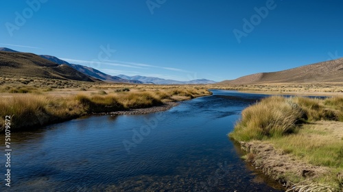 A peaceful stream winding through a mountainous landscape, with a cloudless blue sky as a backdrop.