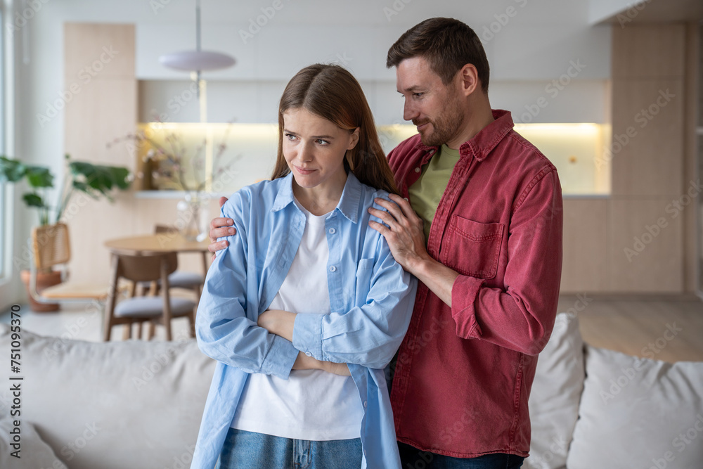 Obraz premium Caring guy gently hugs girlfriend by shoulders trying to look into eyes with opportunity make peace. Husband asks for forgiveness to offended woman showing no emotion at home. Family sincere support.