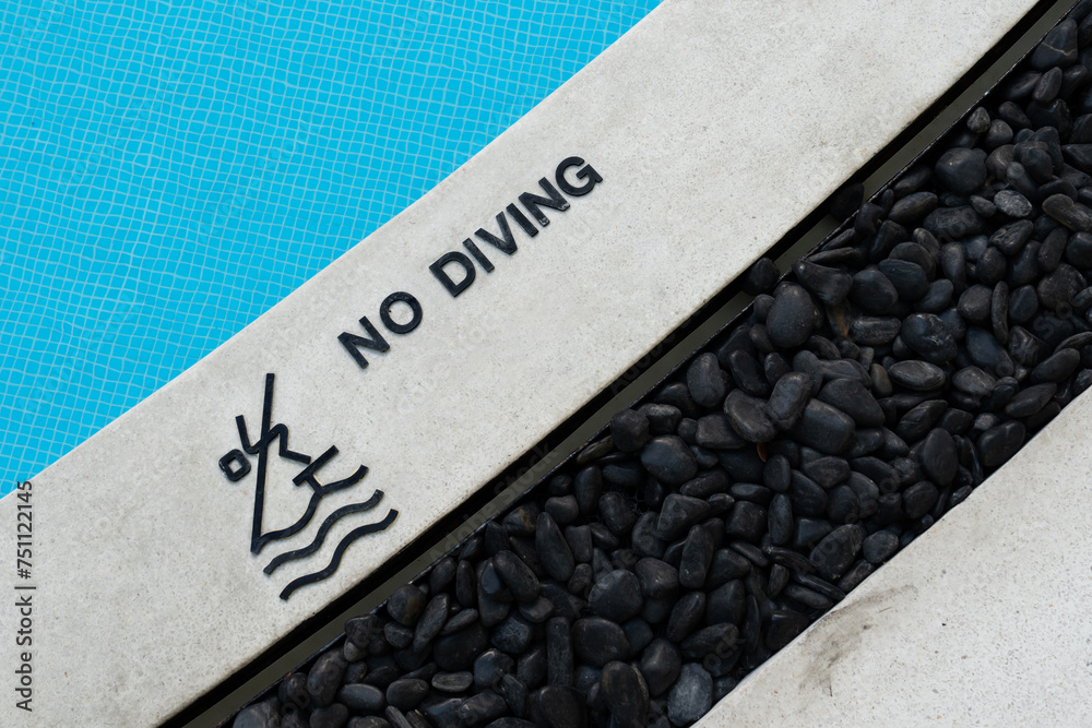 No Diving text warning sign and rules icon at a pool edge with a pictogram beside a blue tiled swimming pool and decorative black stones exterior design No Diving text warning sign and rules icon at a pool edge with a pictogram beside a blue tiled swimming pool and decorative black stones exterior design