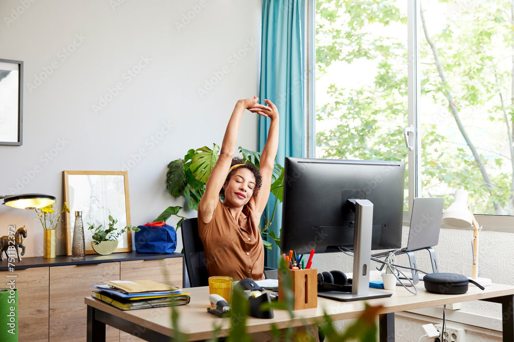 © ALTO IMAGES/Stocksy - Tired woman stretching arms at computer desk in office