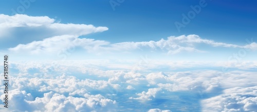 The view from an airplane window shows a clear blue sky with fluffy white clouds scattered below. The perspective captures the vast expanse of the clouds forming patterns against the horizon.