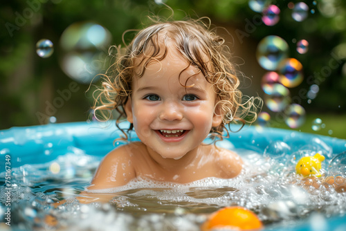 A little cheerful curly girl washes herself in a large basin with toys, a lot of foam and bubbles. © Dzmitry