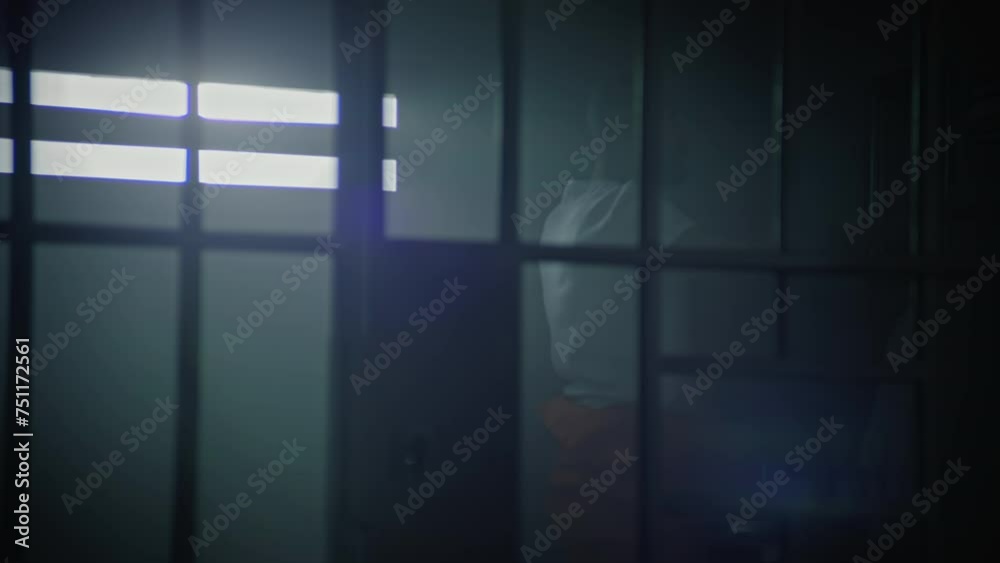 Male prisoner in orange uniform walks around in prison cell, then sits on bed and begins to read Bible. Religious inmate serves imprisonment term in jail. Detention center or correctional facility. Male prisoner in orange uniform walks around in prison cell, then sits on bed and begins to read Bible. Religious inmate serves imprisonment term in jail. Detention center or correctional facility.