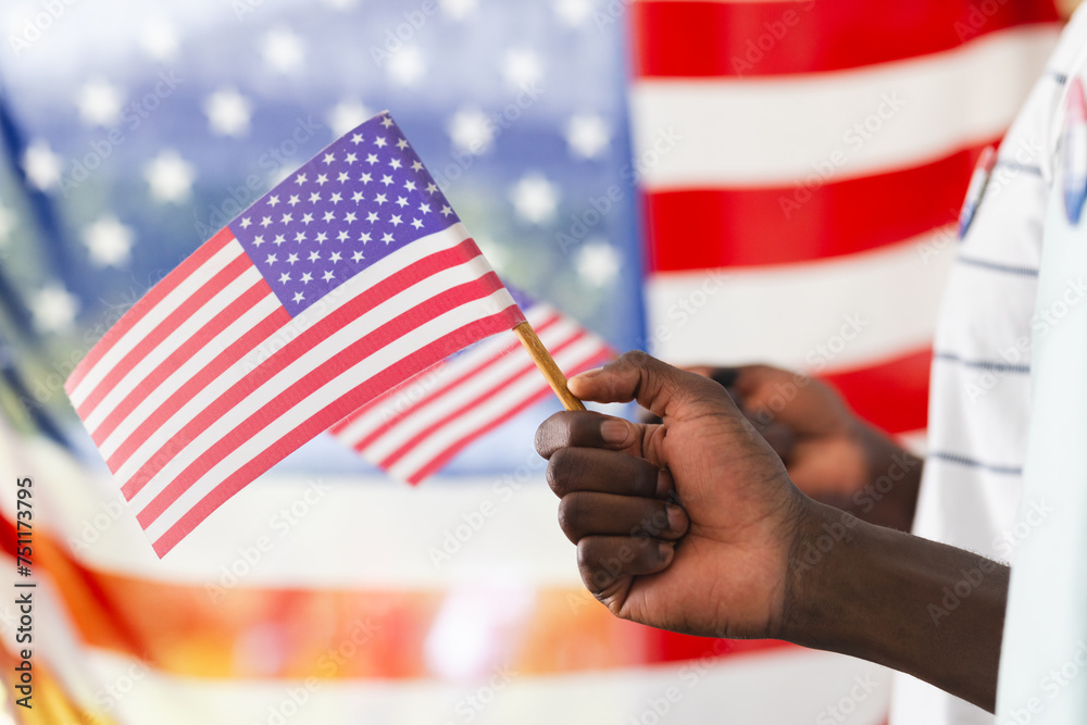 African American man's hand holding a small American flag, with a larger flag in the background