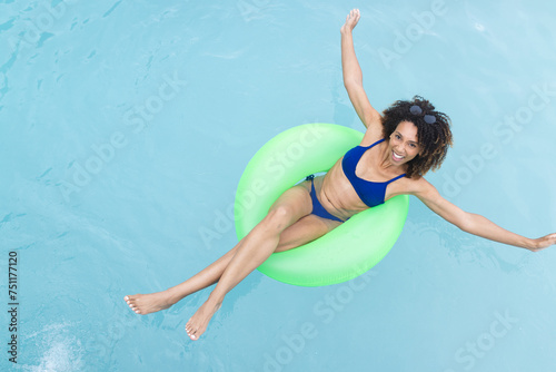 Biracial woman enjoys floating in a pool on a green inner tube, smiling at the camera